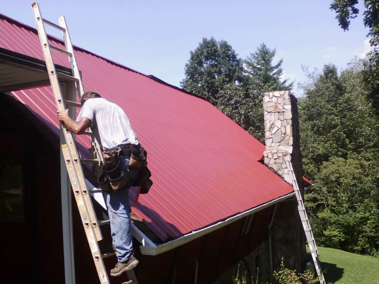 Roofer on a ladder beside a red metal roof and stone chimney — Soffit & Fascia Repair by Roofing Brothers in Canal Fulton